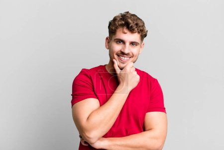Photo for Young adult caucasian man smiling, enjoying life, feeling happy, friendly, satisfied and carefree with hand on chin - Royalty Free Image
