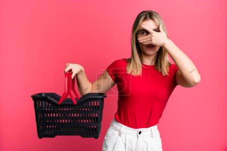 Photo for Young pretty woman looking shocked, scared or terrified, covering face with hand. empty shopping basket concept - Royalty Free Image