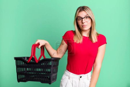 Téléchargez les photos : Young pretty woman shrugging, feeling confused and uncertain. empty shopping basket concept - en image libre de droit