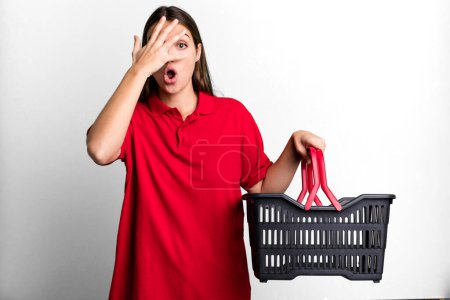 Téléchargez les photos : Young pretty woman looking shocked, scared or terrified, covering face with hand. empty shopping basket concept - en image libre de droit