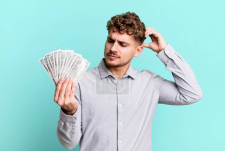 Foto de Young adult caucasian man feeling puzzled and confused, scratching head. dollar bank notes concept - Imagen libre de derechos