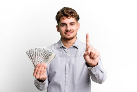 Foto de Young adult caucasian man smiling and looking friendly, showing number one. dollar bank notes concept - Imagen libre de derechos