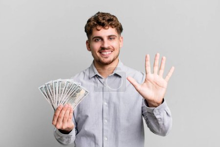 Foto de Young adult caucasian man smiling and looking friendly, showing number five. dollar bank notes concept - Imagen libre de derechos