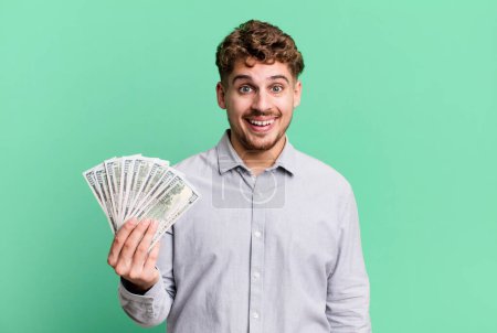 Foto de Young adult caucasian man looking happy and pleasantly surprised. dollar bank notes concept - Imagen libre de derechos