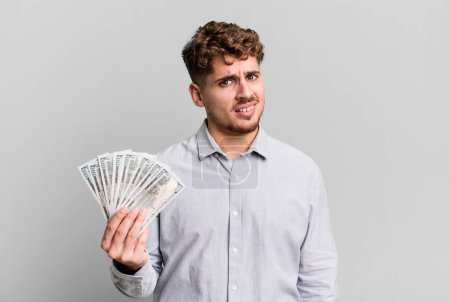 Foto de Young adult caucasian man feeling puzzled and confused. dollar bank notes concept - Imagen libre de derechos