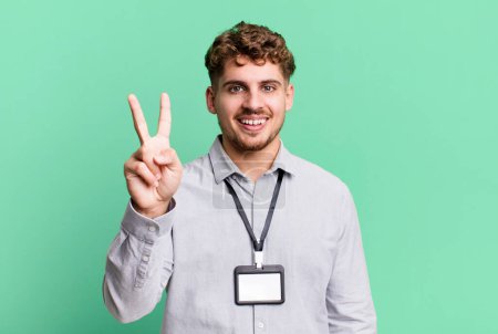 Foto de Young adult caucasian man smiling and looking friendly, showing number two. blank accreditation pass card id concept - Imagen libre de derechos