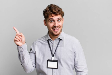 Foto de Young adult caucasian man looking excited and surprised pointing to the side. blank accreditation pass card id concept - Imagen libre de derechos