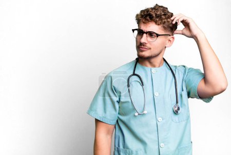 Foto de Young adult caucasian man feeling puzzled and confused, scratching head. nurse concept - Imagen libre de derechos
