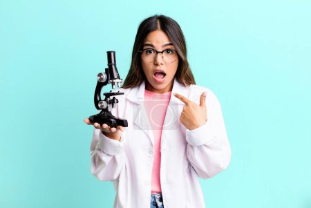 Photo for Hispanic pretty woman looking shocked and surprised with mouth wide open, pointing to self. scients student with a microscope - Royalty Free Image