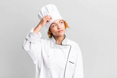 Foto de Cabello rojo bonita mujer sintiéndose desconcertado y confundido, arañando la cabeza. concepto de chef - Imagen libre de derechos