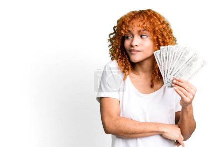 Foto de Cabello rojo mujer bonita encogiéndose de hombros, sintiéndose confundida e incierta. billetes en dólares y concepto de dinero - Imagen libre de derechos