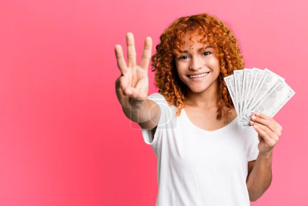 Téléchargez les photos : Red hair pretty woman smiling and looking friendly, showing number three. dollar banknotes and money concept - en image libre de droit