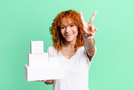 Foto de Red hair pretty woman smiling and looking happy, gesturing victory or peace. blank packages boxes concept - Imagen libre de derechos