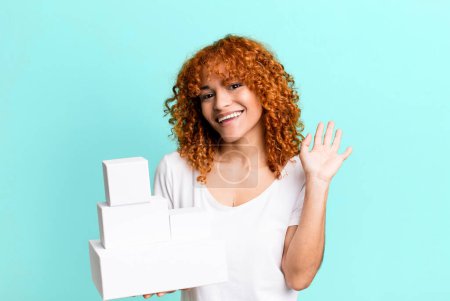 Photo for Red hair pretty woman smiling happily, waving hand, welcoming and greeting you. blank packages boxes concept - Royalty Free Image