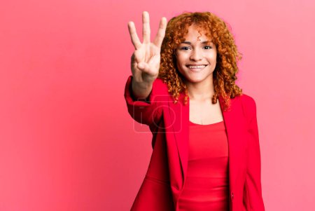 Téléchargez les photos : Cheveux roux jolie femme souriante et sympathique, montrant le numéro trois. concept de femme d'affaires - en image libre de droit