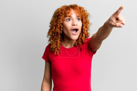 Photo for Redhair pretty woman feeling shocked and surprised, pointing and looking upwards in awe with amazed, open-mouthed look - Royalty Free Image