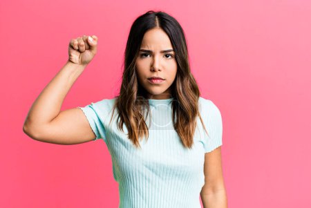 Photo for Hispanic pretty woman feeling serious, strong and rebellious, raising fist up, protesting or fighting for revolution - Royalty Free Image