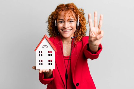 Foto de Pelo rojo bonita mujer agente de bienes raíces con un modelo de casa y auriculares - Imagen libre de derechos
