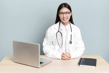 Foto de Bonita mujer asiática sonriendo felizmente con una mano en la cadera y con confianza. escritorio médico y portátil - Imagen libre de derechos