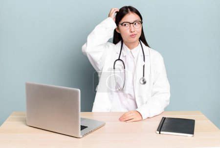 Foto de Pretty asian woman smiling happily and daydreaming or doubting. physician desk and laptop - Imagen libre de derechos