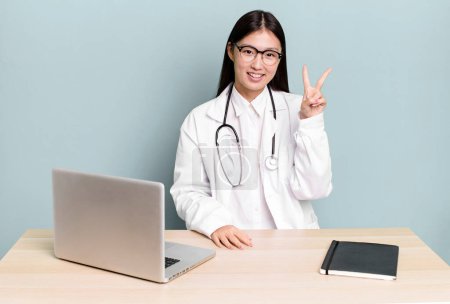 Photo for Pretty asian woman smiling and looking happy, gesturing victory or peace. physician desk and laptop - Royalty Free Image