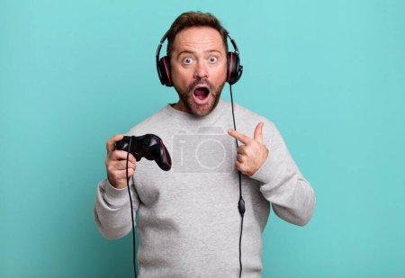 Téléchargez les photos : Homme d'âge moyen se sentant heureux et pointant vers soi-même avec un excité. concept gamer avec un contrôle et un casque - en image libre de droit
