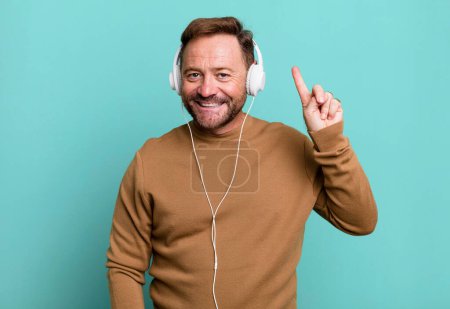 Téléchargez les photos : Homme d'âge moyen souriant et l'air amical, montrant numéro un. écouter de la musique avec un casque - en image libre de droit