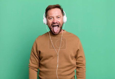 Téléchargez les photos : Homme d'âge moyen se sentant dégoûté et irrité et la langue. écouter de la musique avec un casque - en image libre de droit
