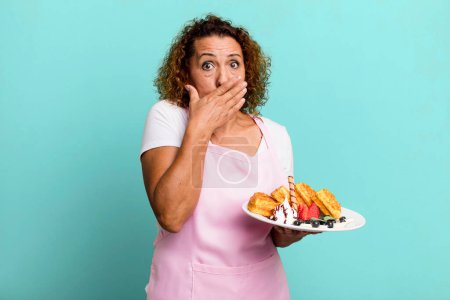 Téléchargez les photos : Pretty middle age woman covering mouth with hands with a shocked. home made waffles concept - en image libre de droit