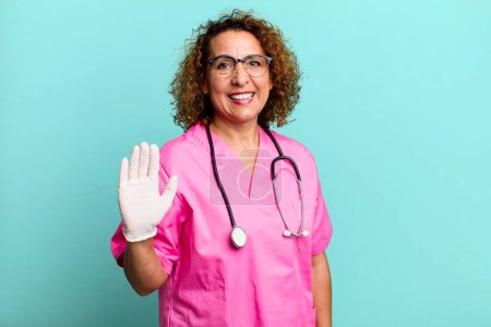 Foto de Pretty middle age woman smiling happily, waving hand, welcoming and greeting you. nurse concept - Imagen libre de derechos