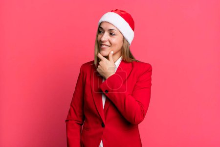 Téléchargez les photos : Pretty blonde woman smiling with a happy, confident expression with hand on chin. christmas and santa hat concept - en image libre de droit