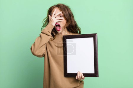 Photo for Hispanic pretty woman looking shocked, scared or terrified, covering face with hand with an empty blank frame - Royalty Free Image