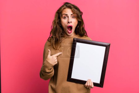 Photo for Hispanic pretty woman looking shocked and surprised with mouth wide open, pointing to self with an empty blank frame - Royalty Free Image