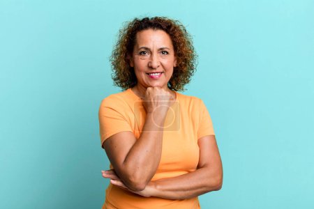 Foto de Mujer hispana de mediana edad mirando feliz y sonriendo con la mano en la barbilla, preguntándose o haciendo una pregunta, comparando opciones - Imagen libre de derechos