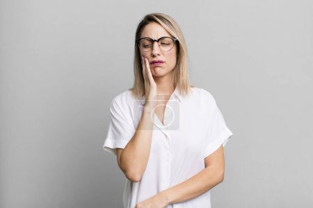 Photo for Blonde adult woman holding cheek and suffering painful toothache, feeling ill, miserable and unhappy, looking for a dentist - Royalty Free Image