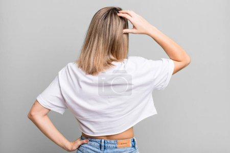 Photo for Blonde adult woman feeling clueless and confused, thinking a solution, with hand on hip and other on head, rear view - Royalty Free Image