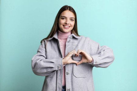 Photo for Pretty young adult woman smiling and feeling happy, cute, romantic and in love, making heart shape with both hands - Royalty Free Image