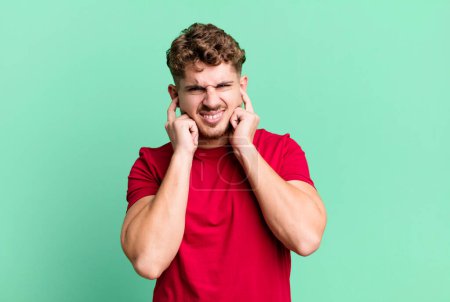 Photo for Young adult caucasian man looking angry, stressed and annoyed, covering both ears to a deafening noise, sound or loud music - Royalty Free Image