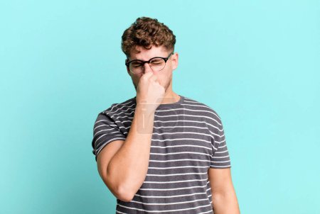 Téléchargez les photos : Young adult caucasian man feeling disgusted, holding nose to avoid smelling a foul and unpleasant stench - en image libre de droit