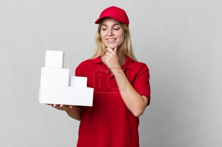Téléchargez les photos : Caucasian blonde woman smiling with a happy, confident expression with hand on chin. delivery boxes - en image libre de droit