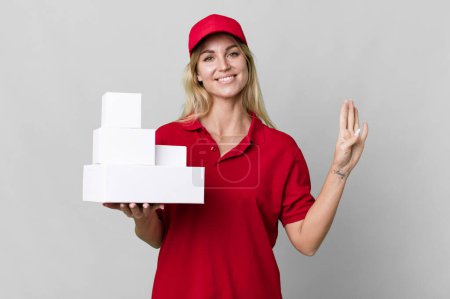 Téléchargez les photos : Caucasian blonde woman smiling and looking friendly, showing number four. delivery boxes - en image libre de droit