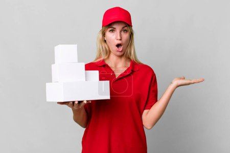 Photo for Caucasian blonde woman looking surprised and shocked, with jaw dropped holding an object. delivery boxes - Royalty Free Image