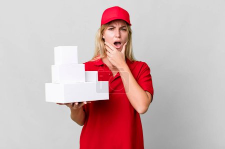 Photo for Caucasian blonde woman with mouth and eyes wide open and hand on chin. delivery boxes - Royalty Free Image