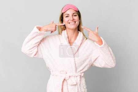 Foto de Caucasian blonde woman smiling confidently pointing to own broad smile. night wear concept - Imagen libre de derechos