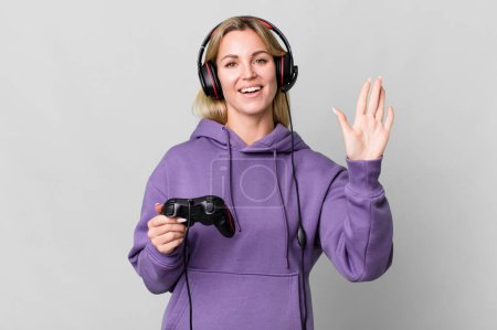 Foto de Caucasian blonde woman smiling happily, waving hand, welcoming and greeting you. gamer concept - Imagen libre de derechos