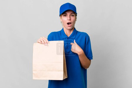Photo for Caucasian blonde woman looking shocked and surprised with mouth wide open, pointing to self. paper bag delivery concept - Royalty Free Image