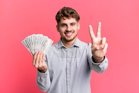Foto de Young adult caucasian man smiling and looking happy, gesturing victory or peace. dollar bank notes concept - Imagen libre de derechos