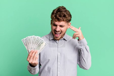 Foto de Young adult caucasian man looking unhappy and stressed, suicide gesture making gun sign. dollar bank notes concept - Imagen libre de derechos