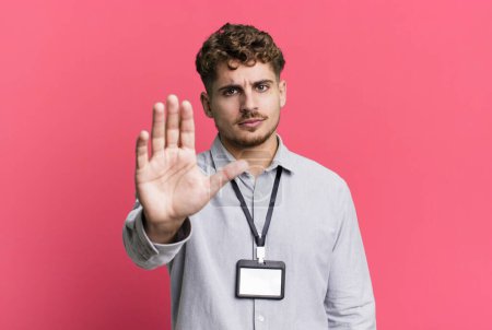 Foto de Young adult caucasian man looking serious showing open palm making stop gesture. blank accreditation pass card id concept - Imagen libre de derechos