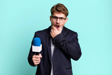Foto de Young adult caucasian man with mouth and eyes wide open and hand on chin. journalist or presenter with a microphone - Imagen libre de derechos